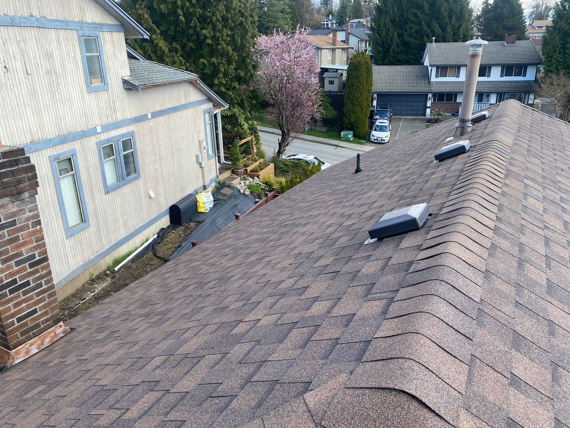 A high-angle view of a brown asphalt shingled roof, showing a chimney, roof vents, and a neighboring house in the background.