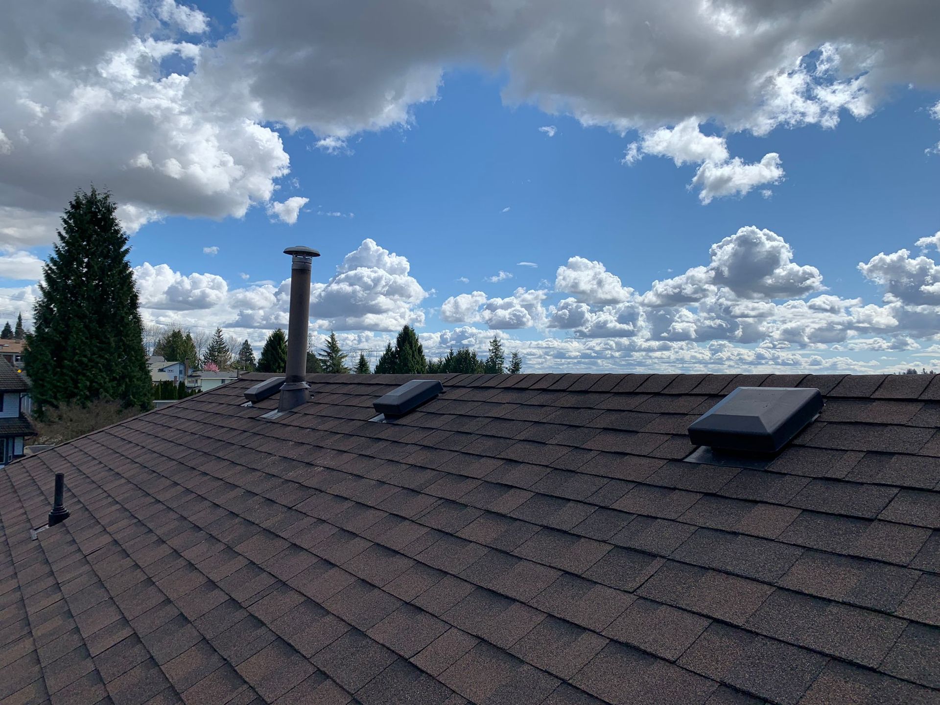 A view of a brown shingled roof with a chimney and three dark roof vents against a bright blue sky with white clouds.
