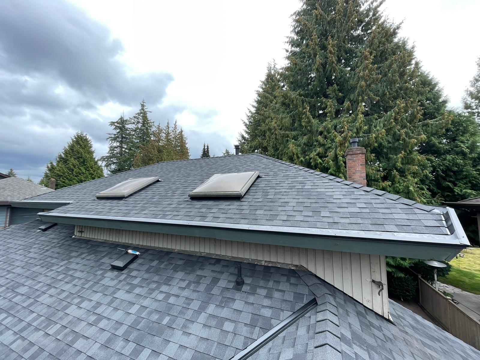 Gray shingled roof with two skylights, chimney, and surrounding trees under cloudy sky.