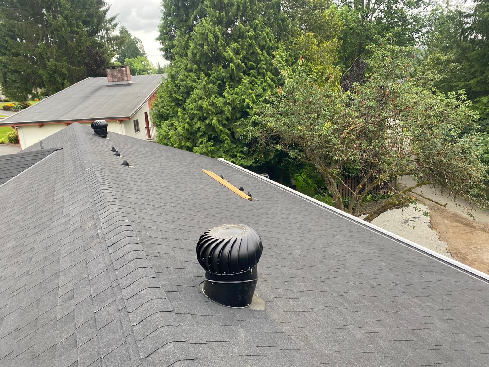 A black wind turbine roof vent centered on a gray shingled roof with trees and another house in the background.