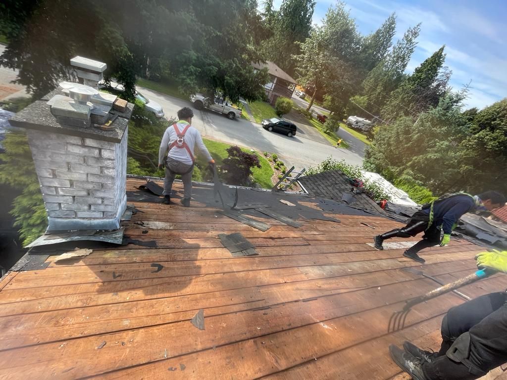 Workers in safety harnesses remove shingles from a wooden residential roof next to a white brick chimney.