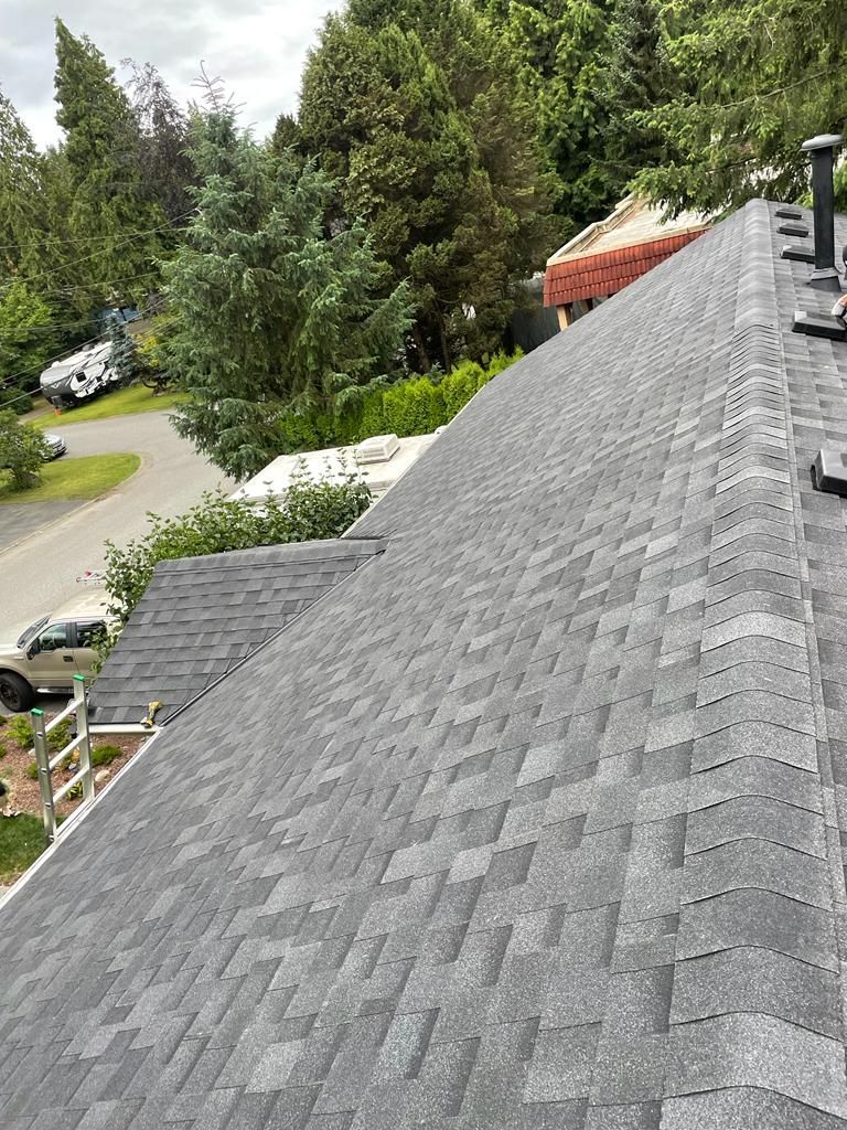 A high-angle view of a newly installed dark grey asphalt shingle roof on a house, looking down toward a street and trees.