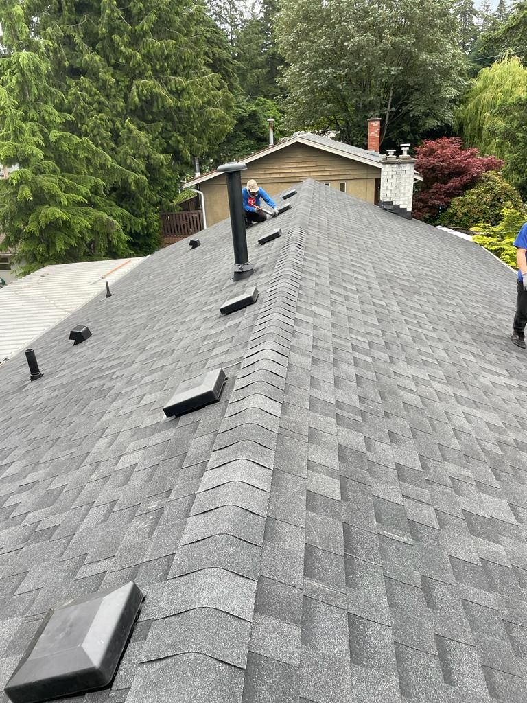 Workers on a grey shingled roof with vents and a chimney, surrounded by trees on a sunny day.