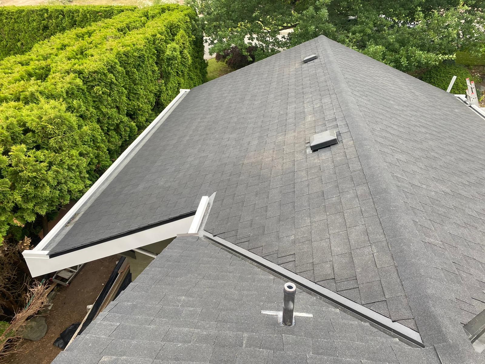 A high-angle view of a grey shingled roof with a vent pipe and a gutter system, bordered by a tall, green hedge.