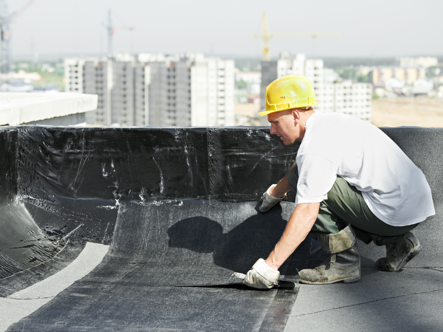A construction worker in a hard hat applies black roofing material to a flat roof on a sunny day.