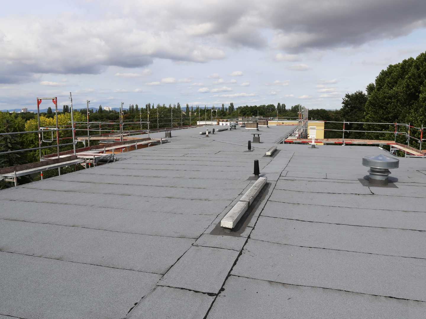 Flat rooftop with gray membrane, vents, and safety railings under a cloudy sky.