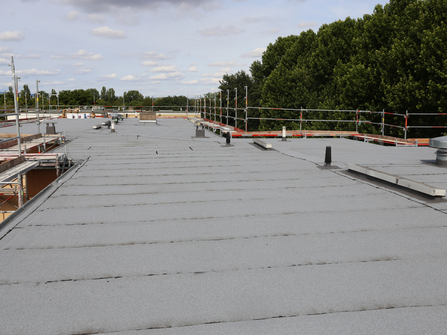 A flat grey roof under construction, featuring scaffolding along the edges and several pipes protruding from the surface.
