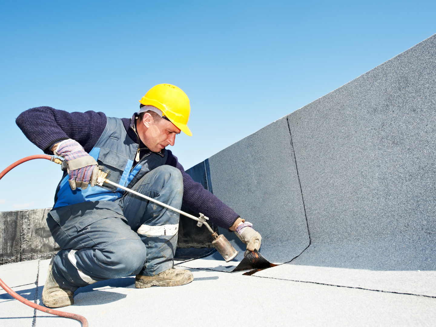 A worker in a yellow hard hat and work clothes uses a propane torch to seal roofing material on a flat roof.