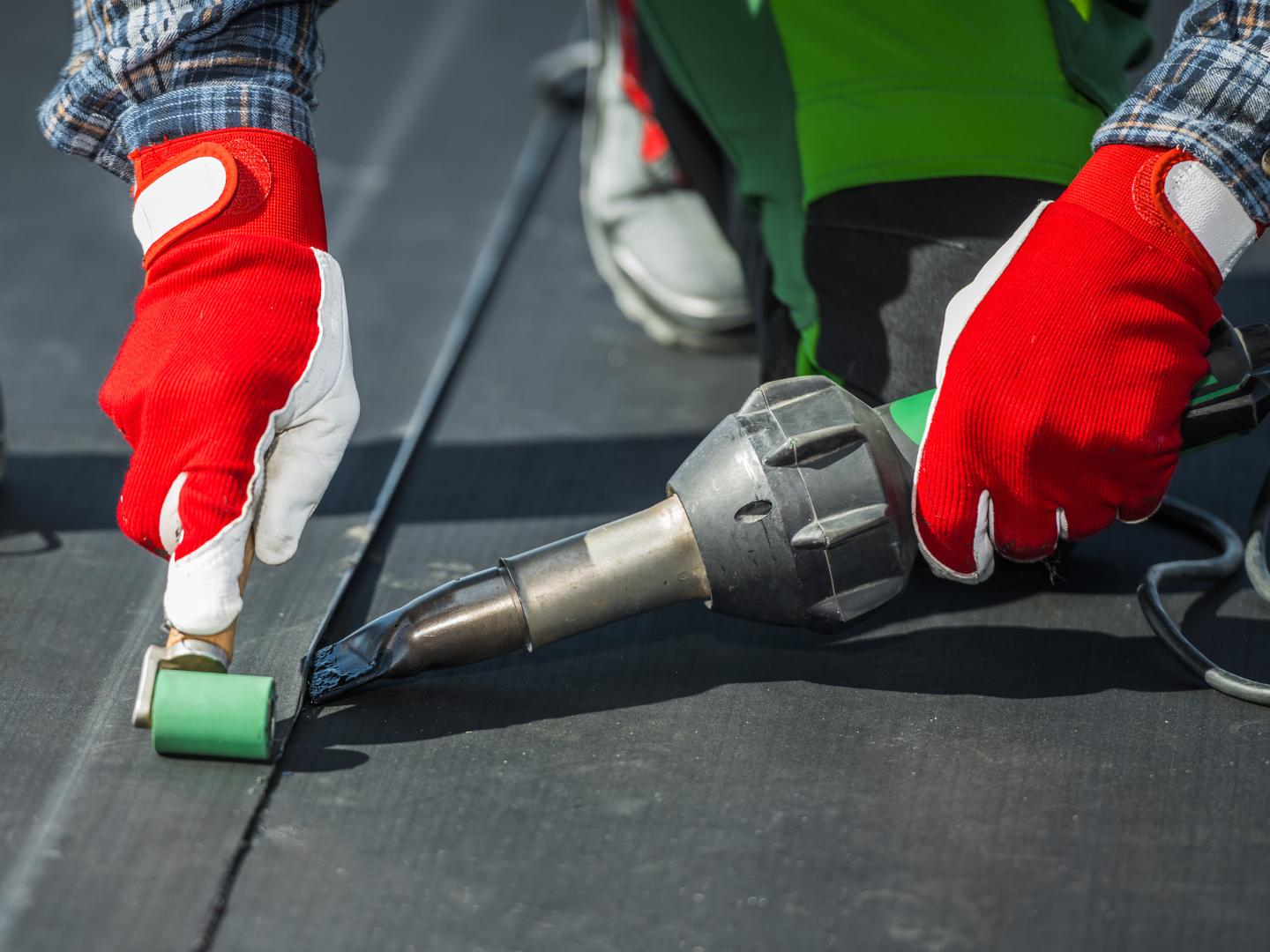 A worker in red gloves uses a hot-air welding tool and a green roller to seal a seam on black roofing material.