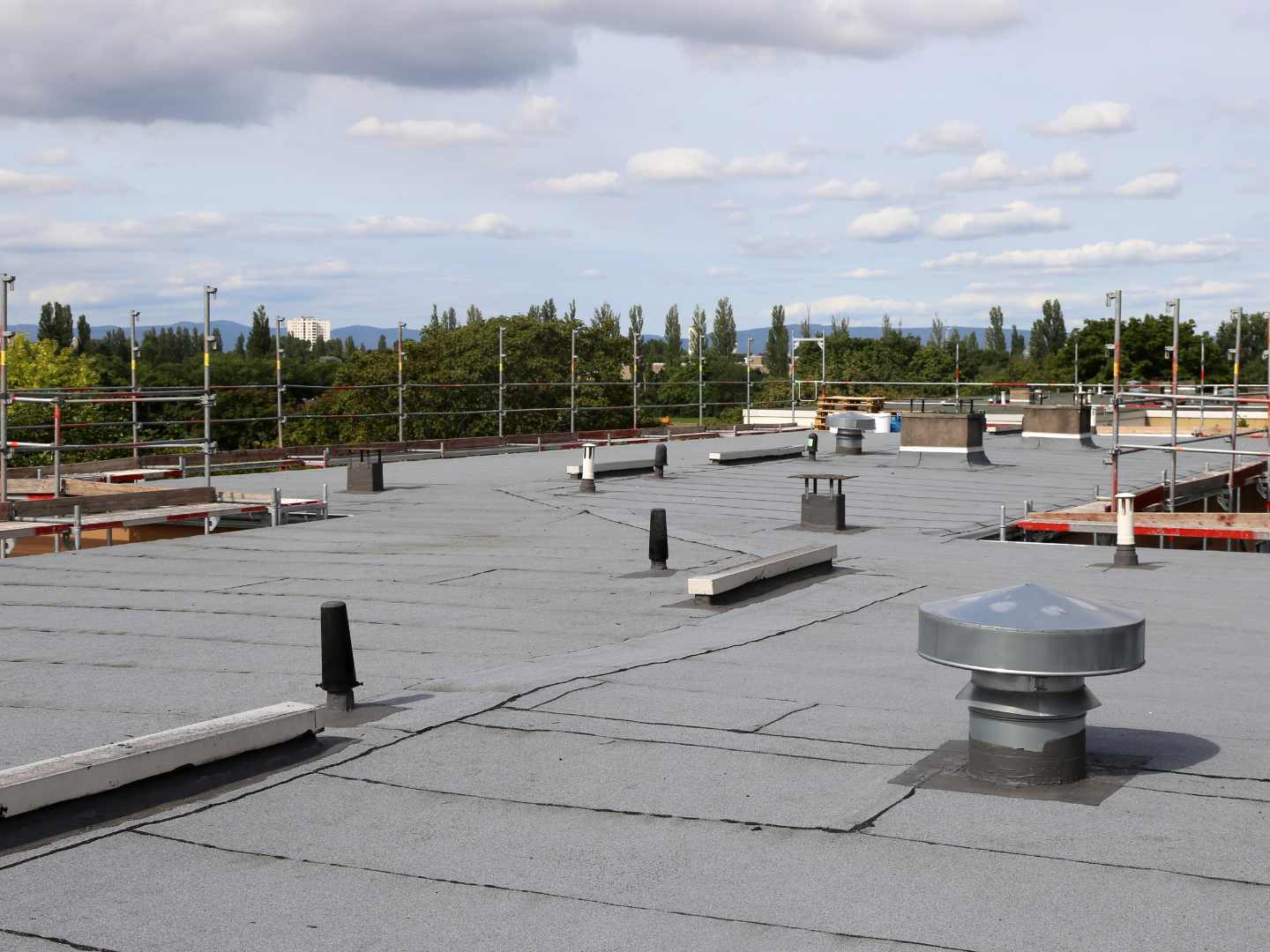 A flat, grey industrial roof featuring scattered ventilation pipes, a metal vent cap, and perimeter safety scaffolding.