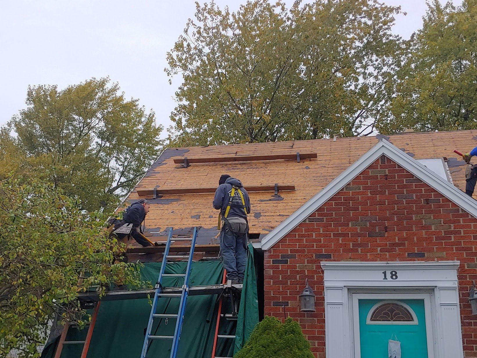 Two people working on the roof of a red brick house with green tarps covering the front below.