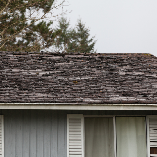 A house roof with severely weathered, curled, and deteriorated dark gray shingles.