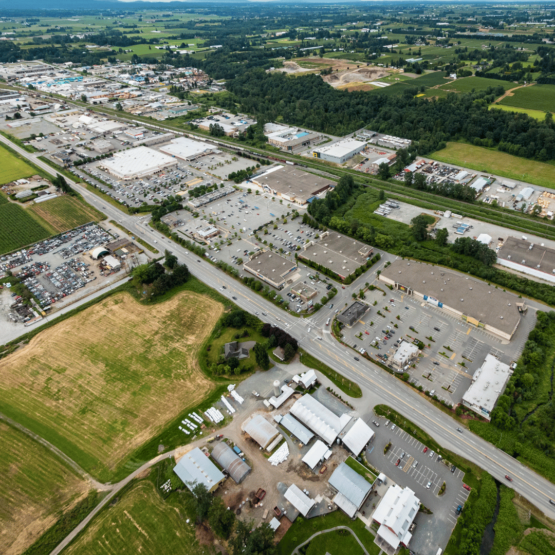 An aerial view of commercial warehouses and parking lots bordering a road, surrounded by fields and patches of trees.