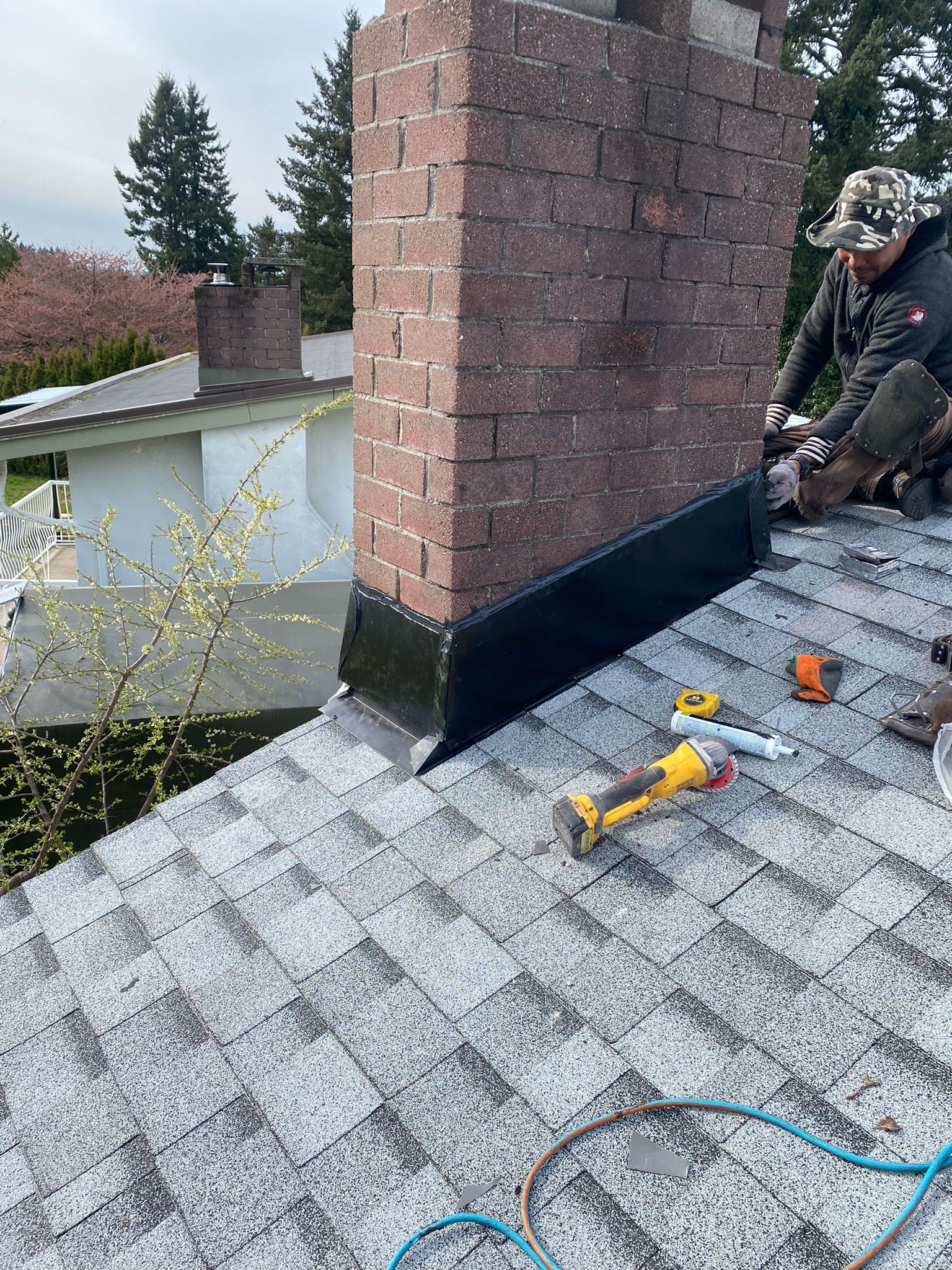 A roofer installs black flashing around a brick chimney on a shingled roof, with tools resting nearby.