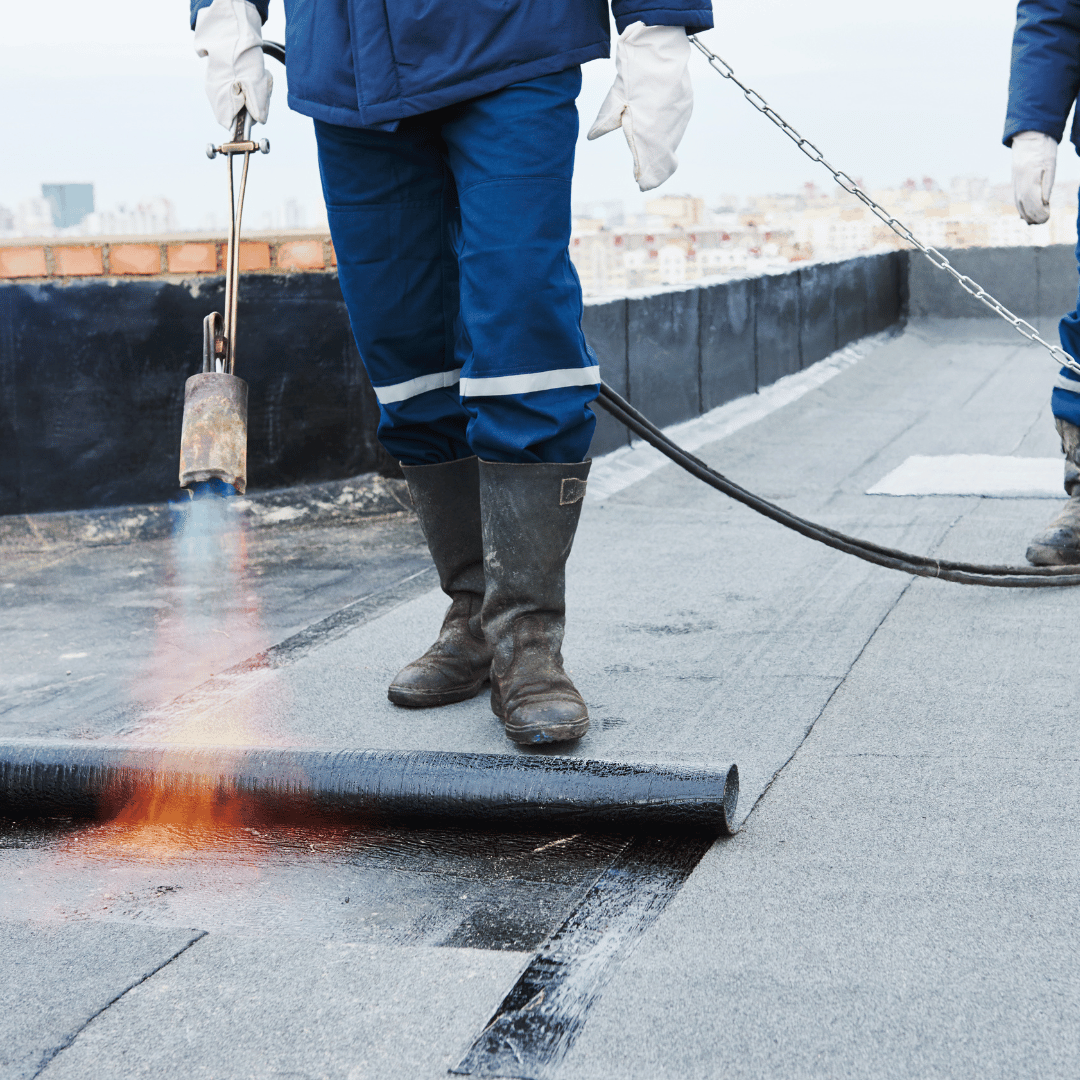 A worker in blue workwear uses a propane torch to heat and apply bitumen roofing felt to a flat roof.