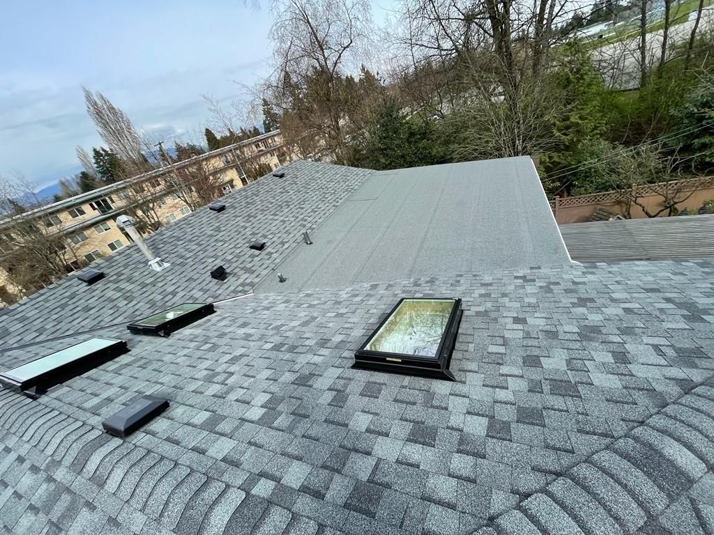 A high-angle view of a grey shingled roof with three rectangular skylights and a section of smooth, flat roofing.