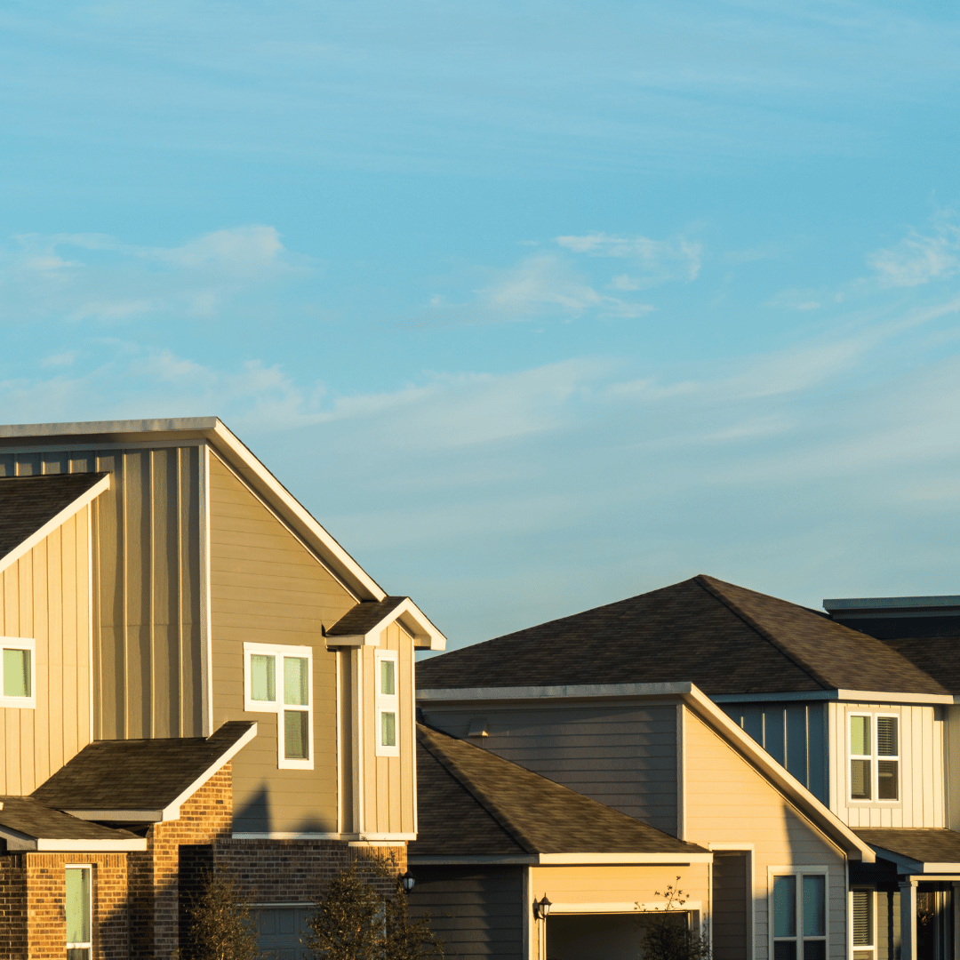 Suburban houses with light tan siding and brown shingled roofs under a clear blue sky.