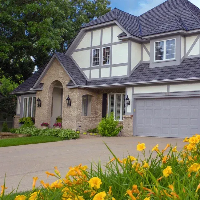 A two-story Tudor-style home with light stucco, stone accents, and a grey garage, framed by yellow daylilies in the front.