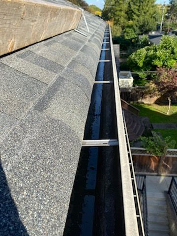 High-angle view of a grey shingled roof edge with metal gutter brackets and a downspout trough overlooking a backyard.
