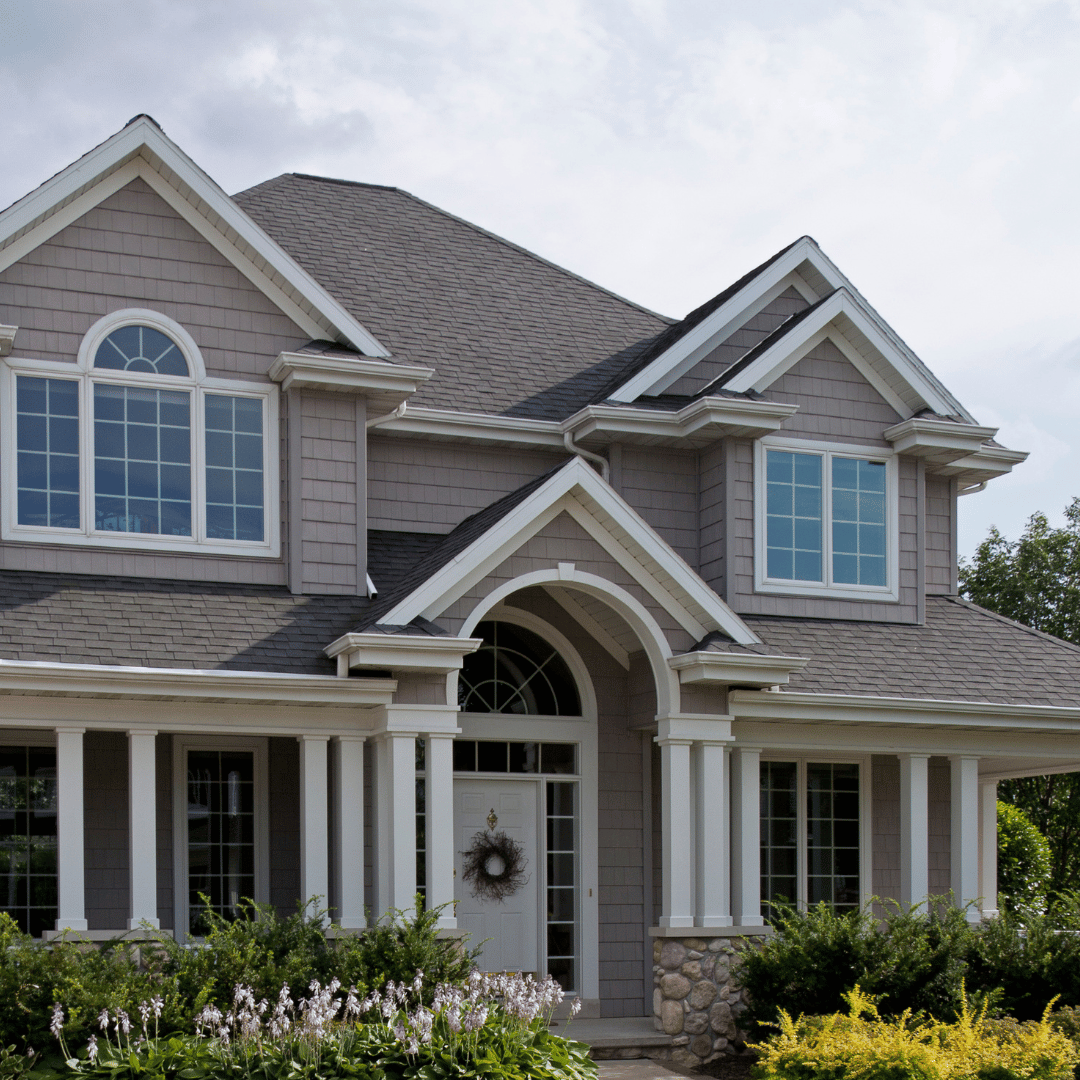 A two-story grey suburban house with white columns, gabled roofs, and a arched entryway with a wreath on the front door.