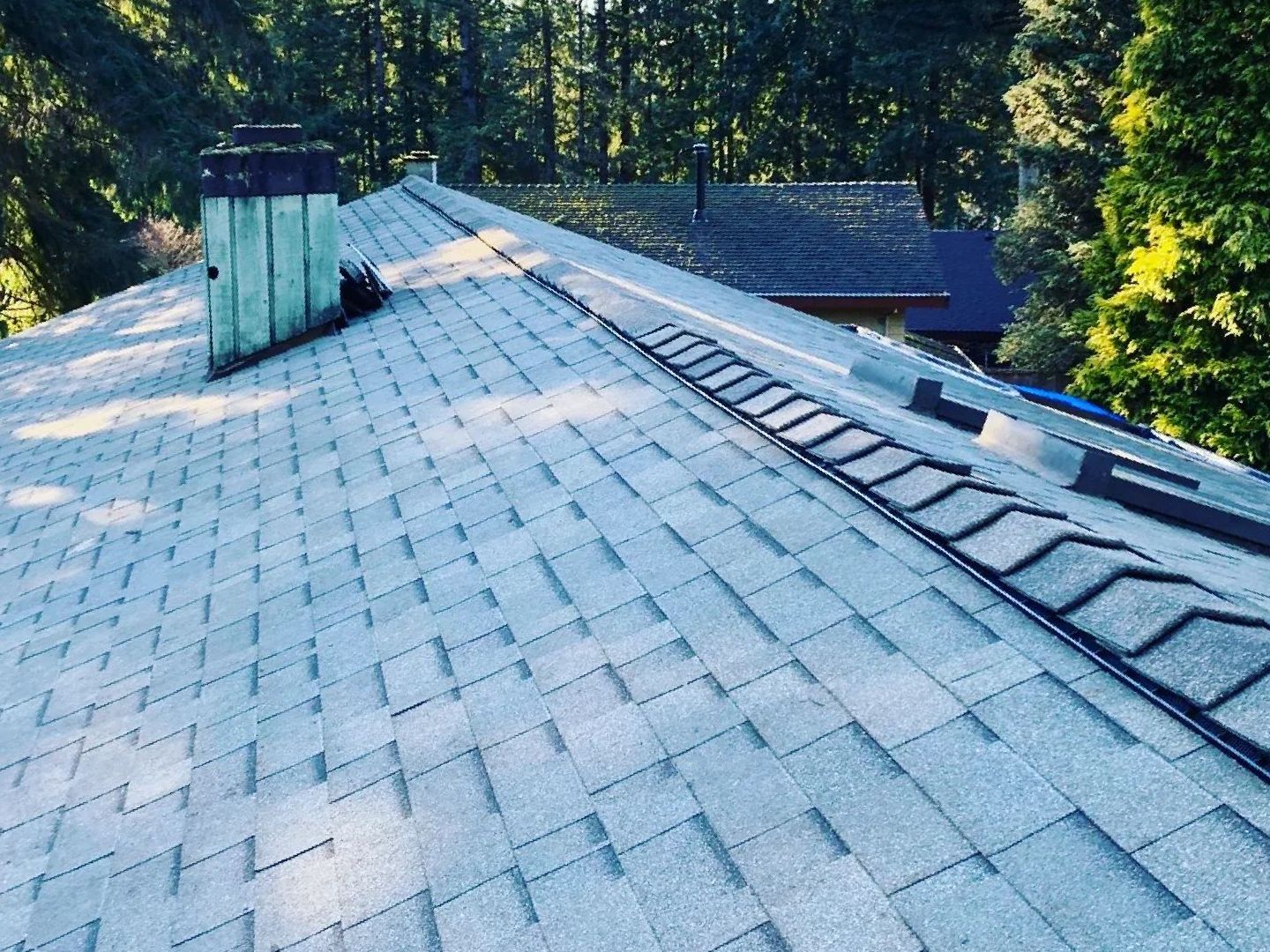 A gray shingled roof with a chimney and a ridge vent, viewed from above, with trees in the background.