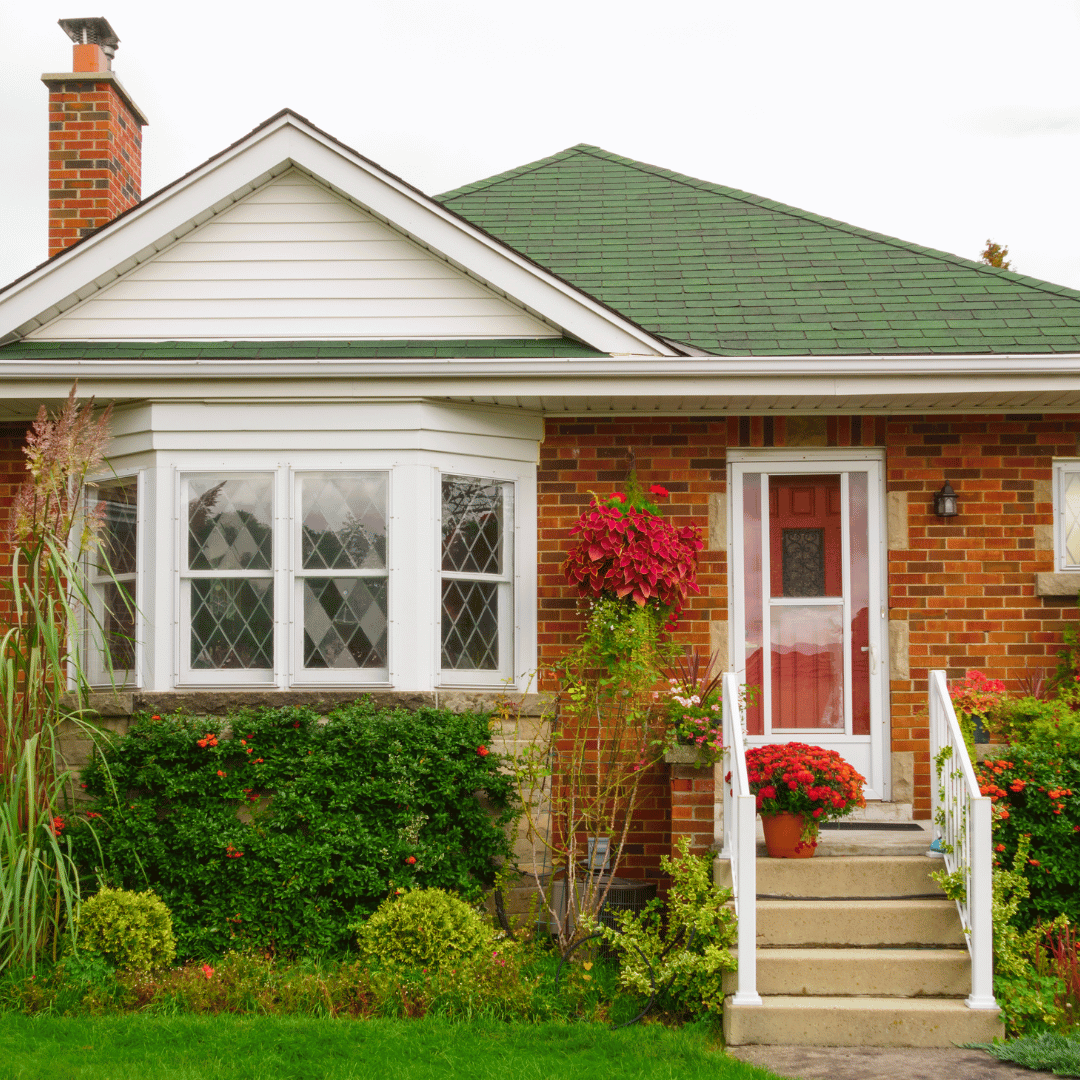 A brick house with a green roof, a large bay window, and a front porch with steps and potted flowers.