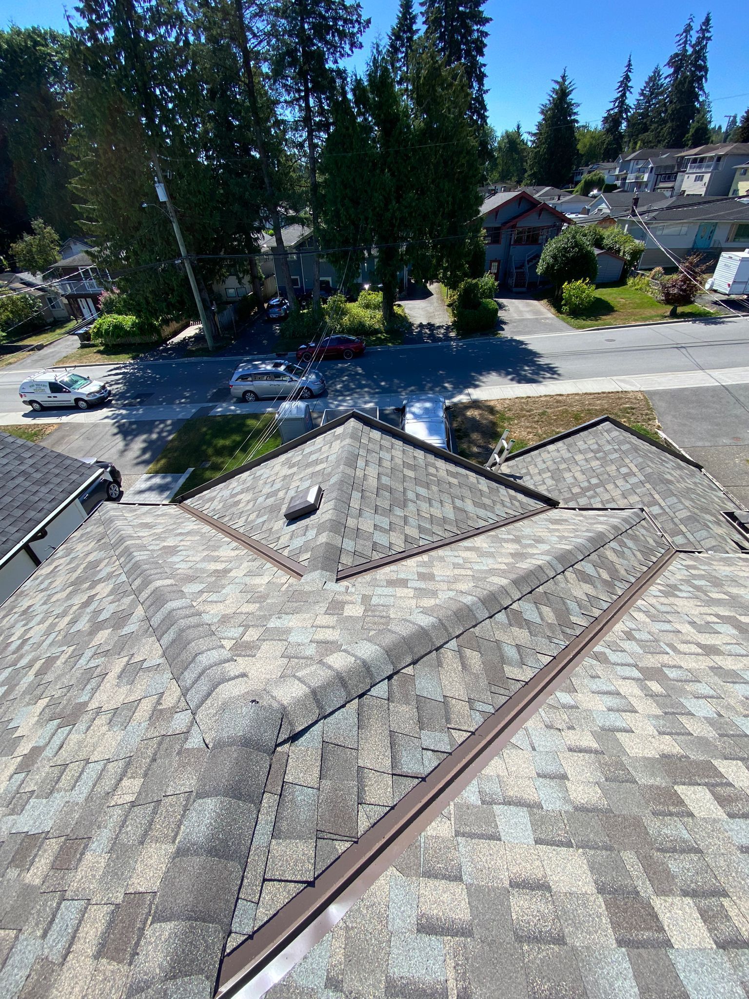 A high-angle view looking down at a gray asphalt shingle roof on a sunny day, with a suburban neighborhood in the background.