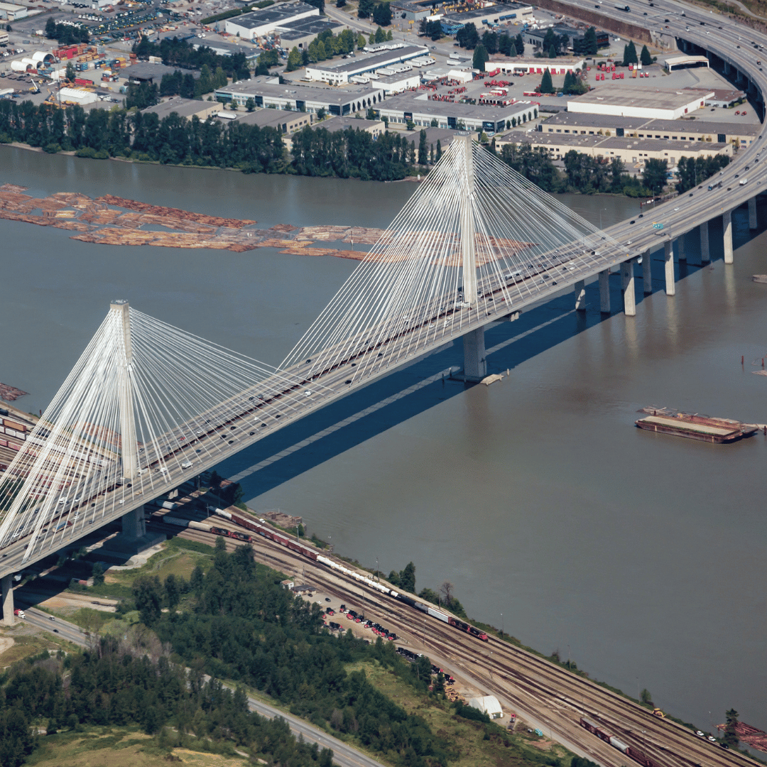 An aerial view of a cable-stayed bridge crossing a wide river near an industrial area and railroad tracks.