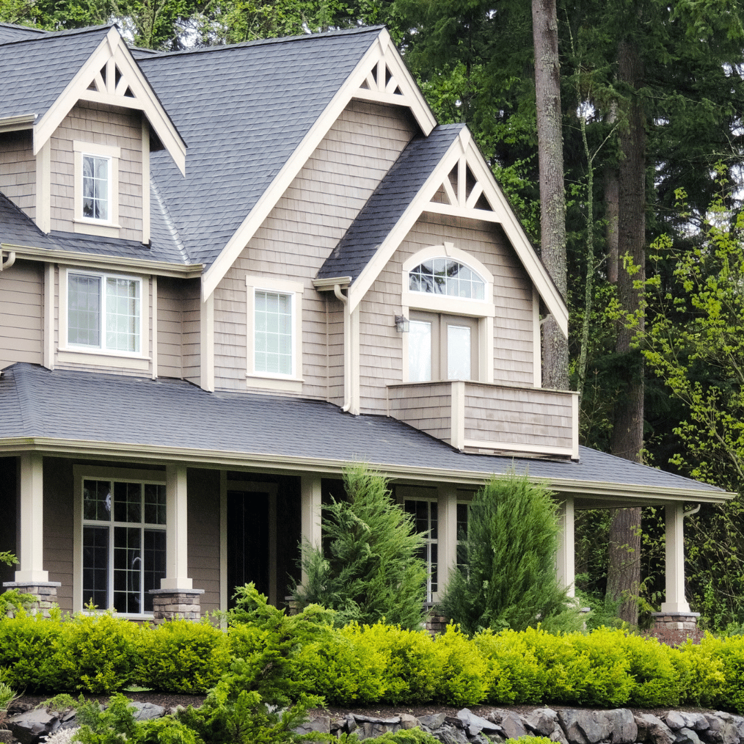 A two-story tan home with shingled siding, a dark gray roof, and a front porch, nestled among lush green trees.
