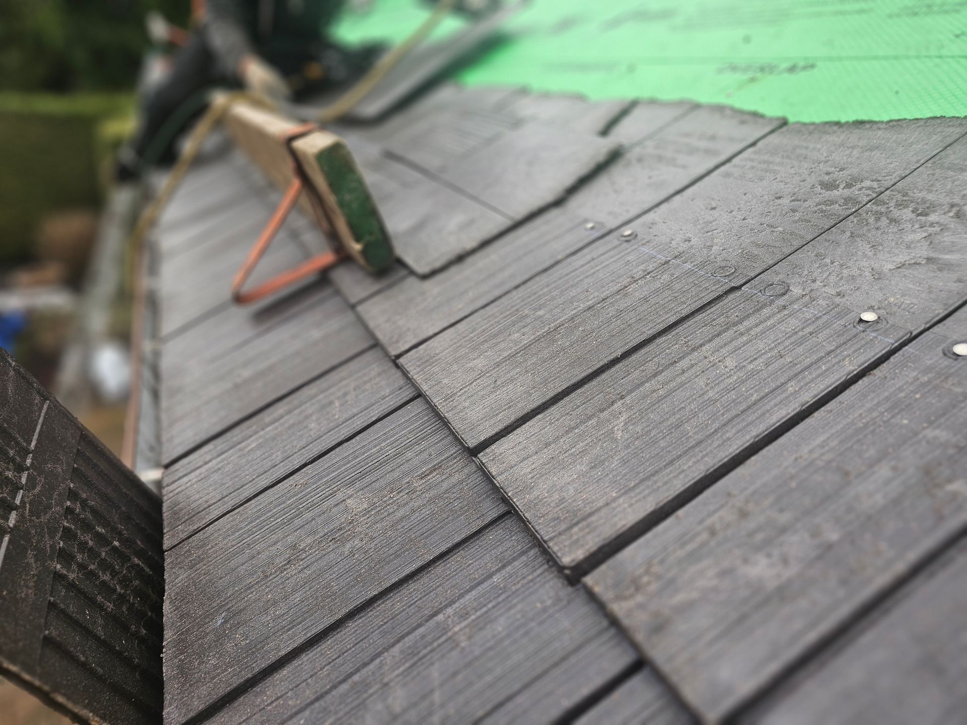 A partially installed grey shingle roof on a house, featuring a wooden roof bracket and a green underlayment strip.