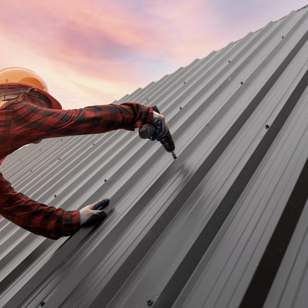 Roofer in orange hardhat screws metal panel to a roof under a sunset sky.