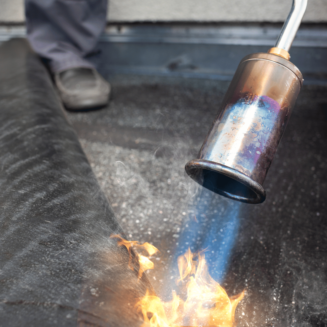 A worker uses a propane torch to melt and seal a roll of black roofing material onto a roof.