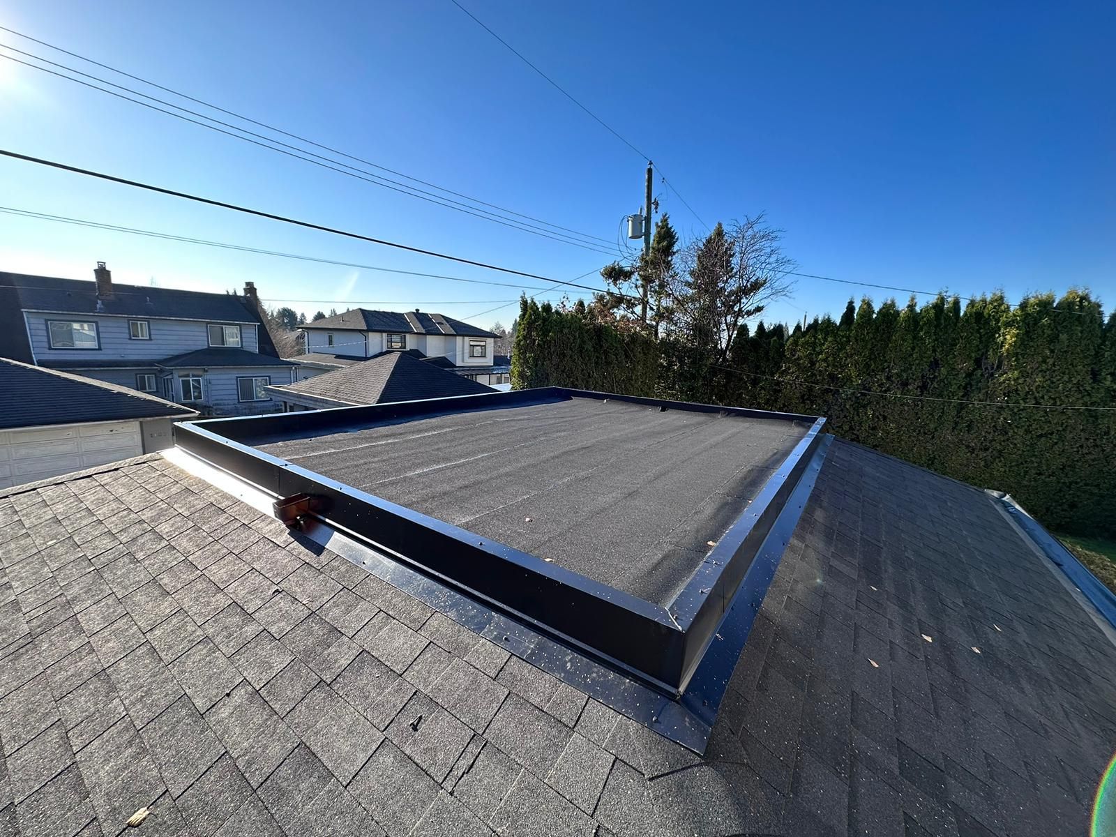 A flat, dark, rubber-membrane roof section surrounded by sloped asphalt shingles under a clear blue sky.