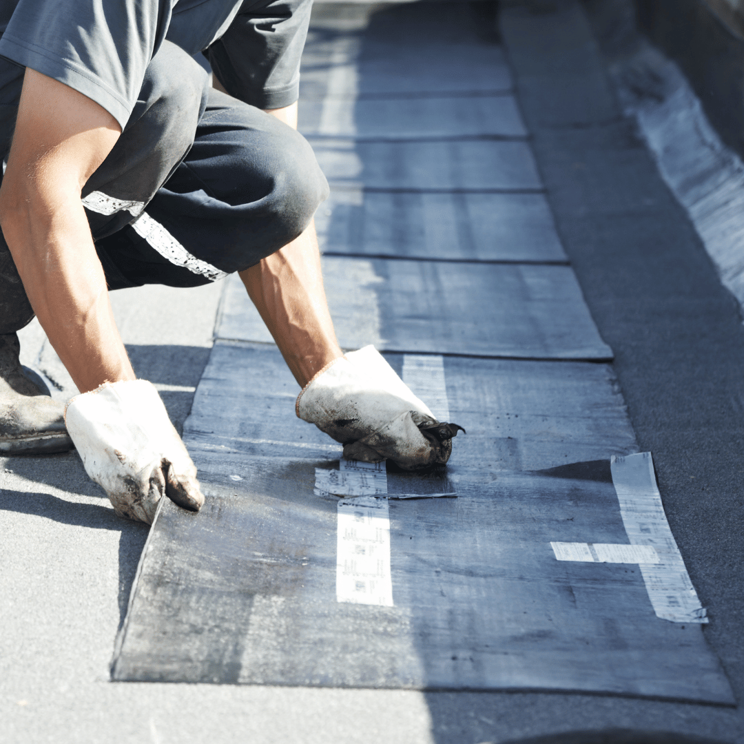 A person in work clothes and gloves installs black roofing membrane material on a flat roof.