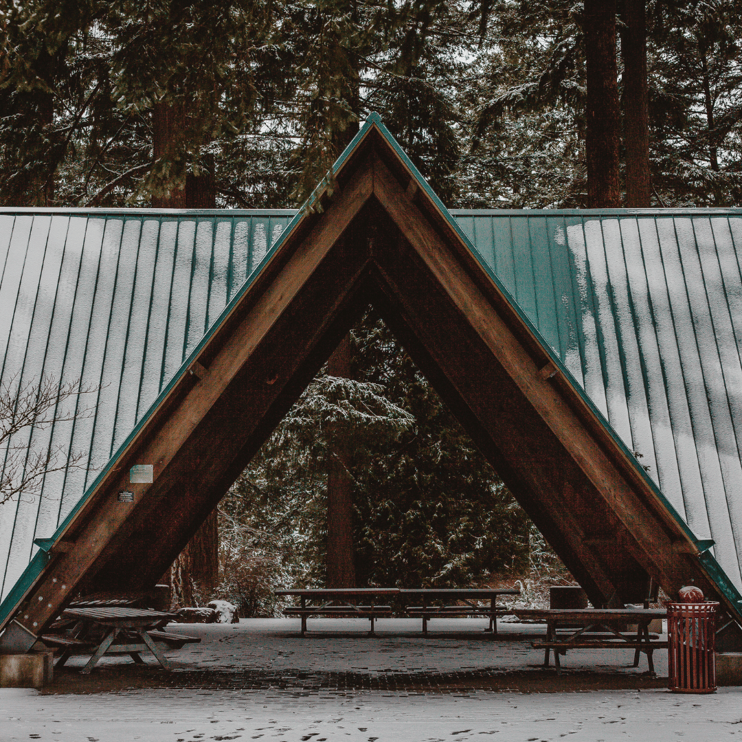 A large, A-frame wooden shelter with a teal metal roof, set in a snowy forest with picnic tables underneath.