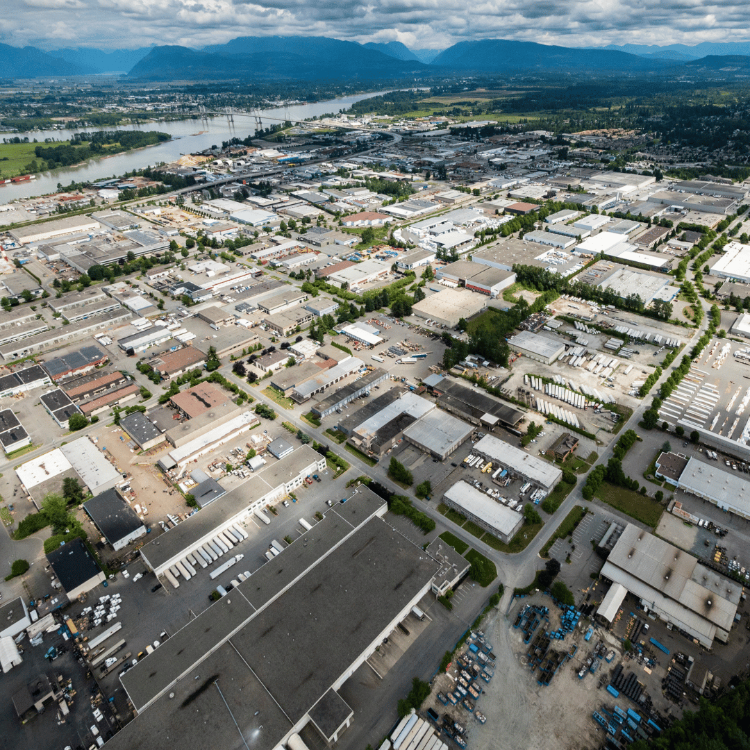 Aerial view of a sprawling industrial district with warehouse buildings and parking lots near a wide river and mountains.