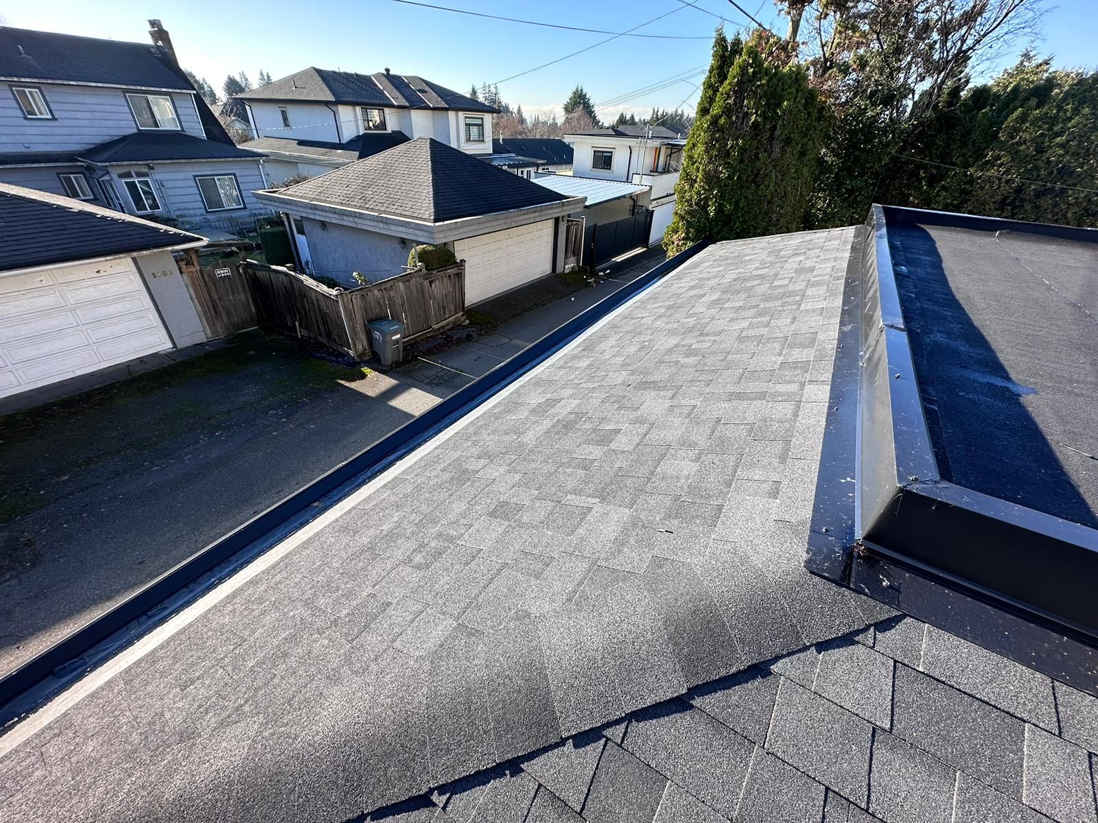 A high-angle view of a grey shingled sloped roof meeting a flat, dark membrane roof section, overlooking a residential alley.