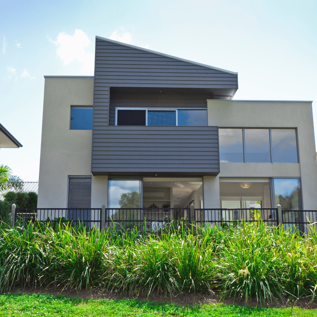 A modern two-story home with a mix of beige stucco and dark horizontal siding, featuring large windows and a front fence.