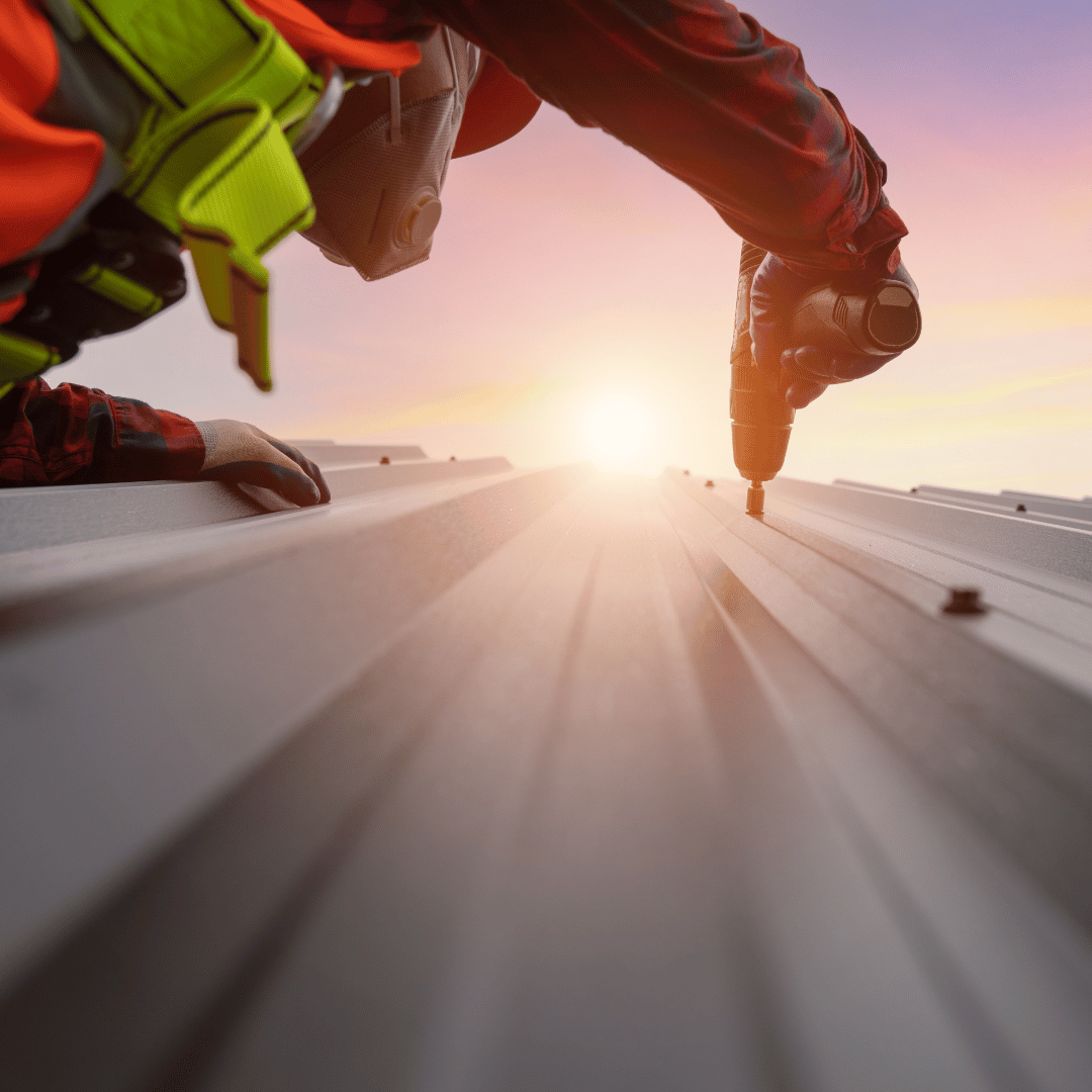 A worker in high-visibility gear uses a drill to secure metal roofing panels during a sunset.