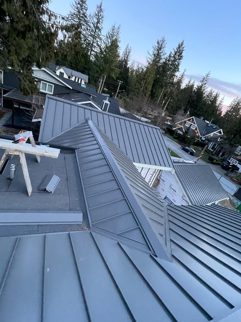 A grey metal roof under construction with a sawhorse and tool set on a flat, tarped area, surrounded by evergreen trees.