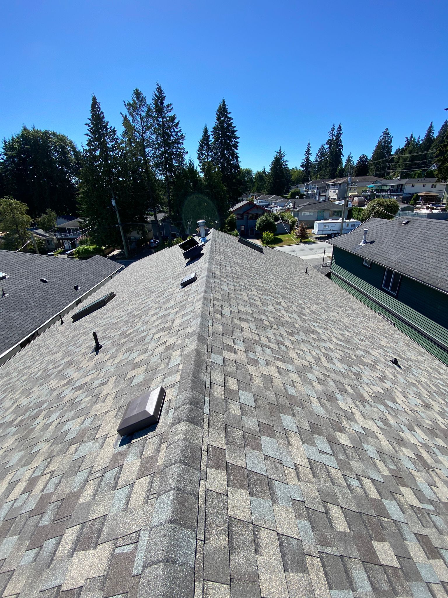 A high-angle view looking down the peaked ridge of a gray asphalt shingle roof under a bright blue sky.