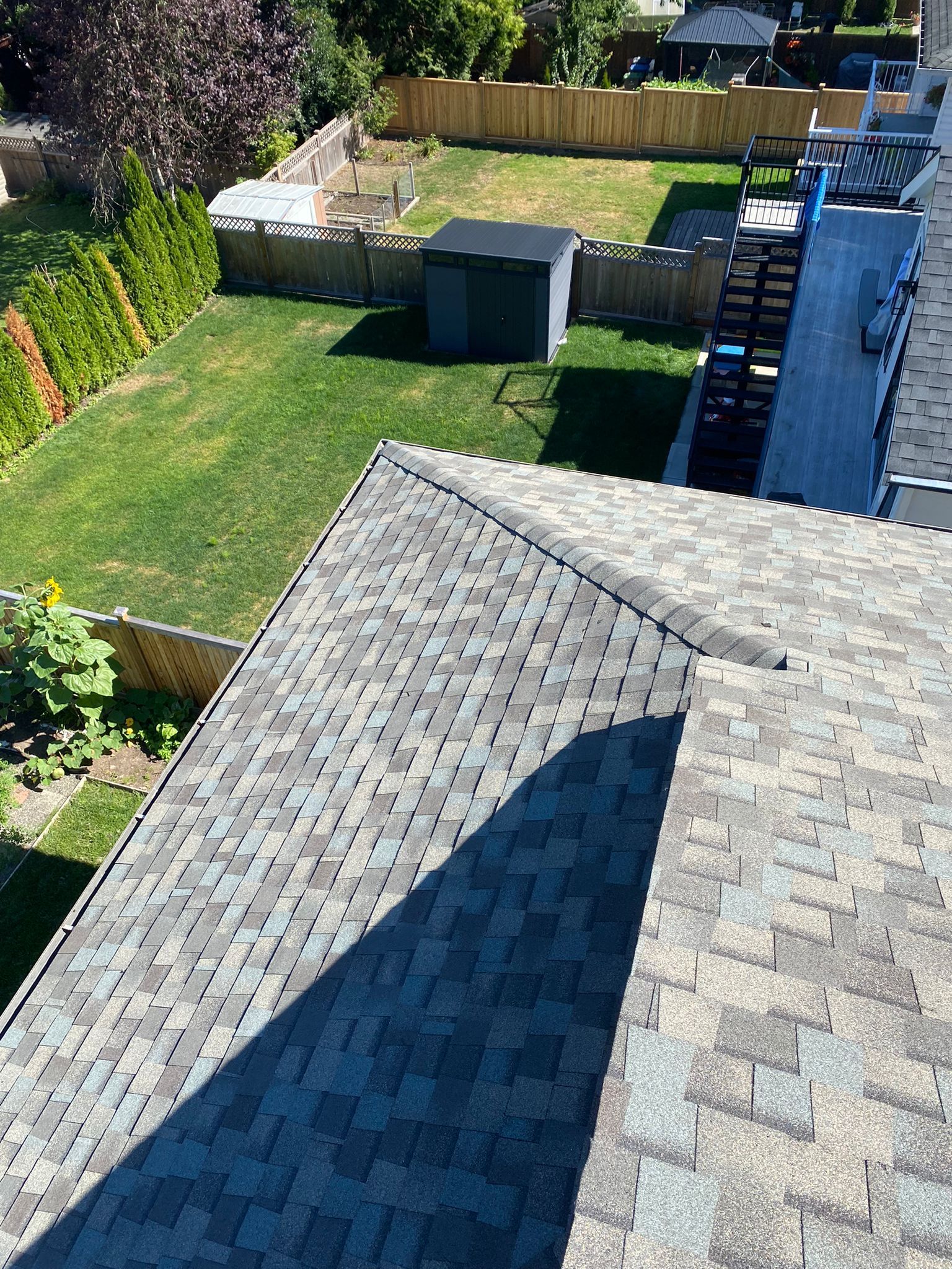 High-angle view of a residential shingled roof overlooking a green backyard with a small shed and wooden fencing.