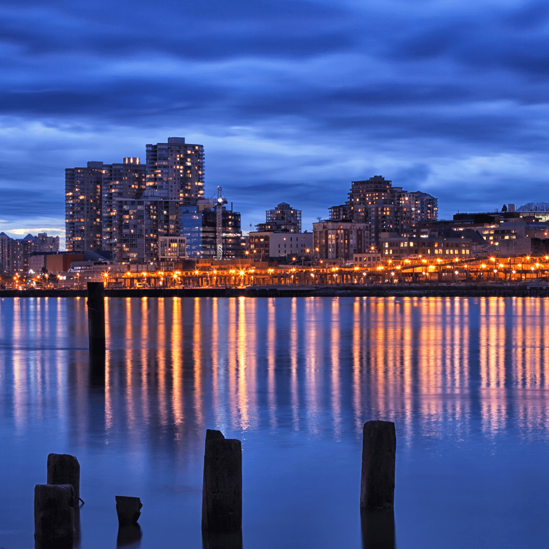 City buildings and lights reflected in a calm body of water at twilight, with wooden pilings in the foreground.