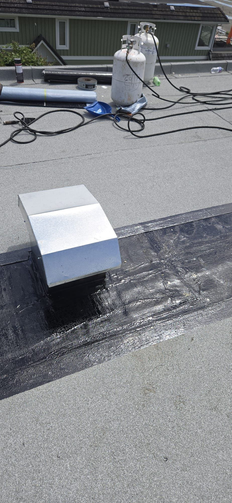 Metal hooded vent beside an exposed membrane strip, with roofing rolls and equipment in the background.