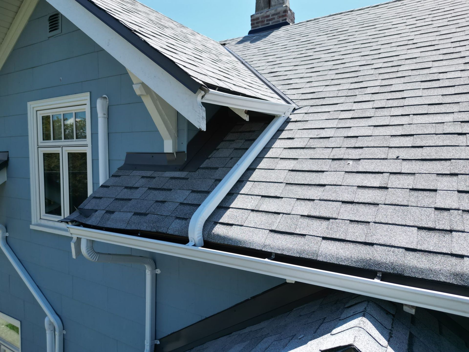 White gutter and downspout routing from a dormer roof into a lower gutter beside blue siding
