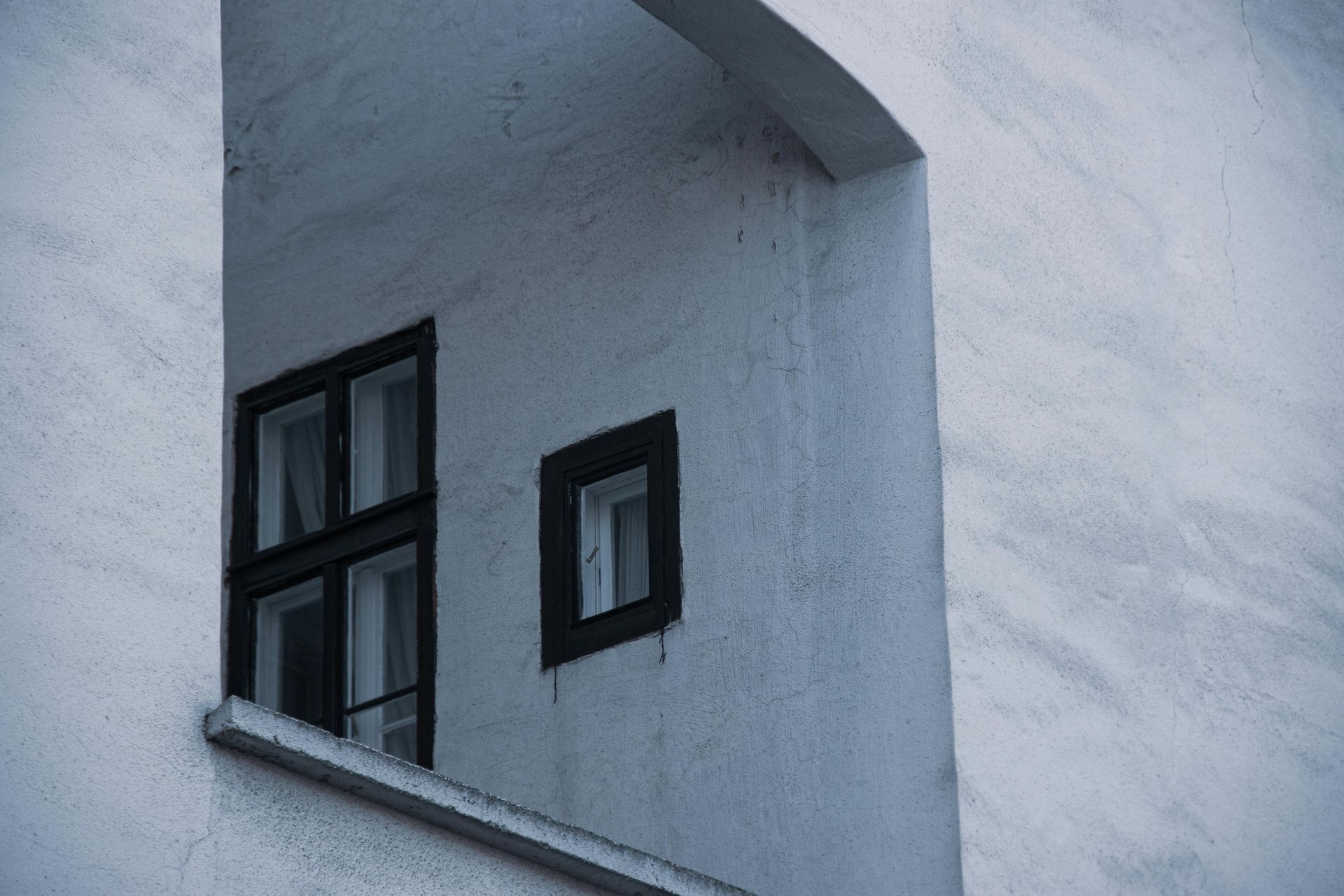 A dark-framed, multi-pane window and a smaller, square window set into a deep, recessed, light gray stone wall.