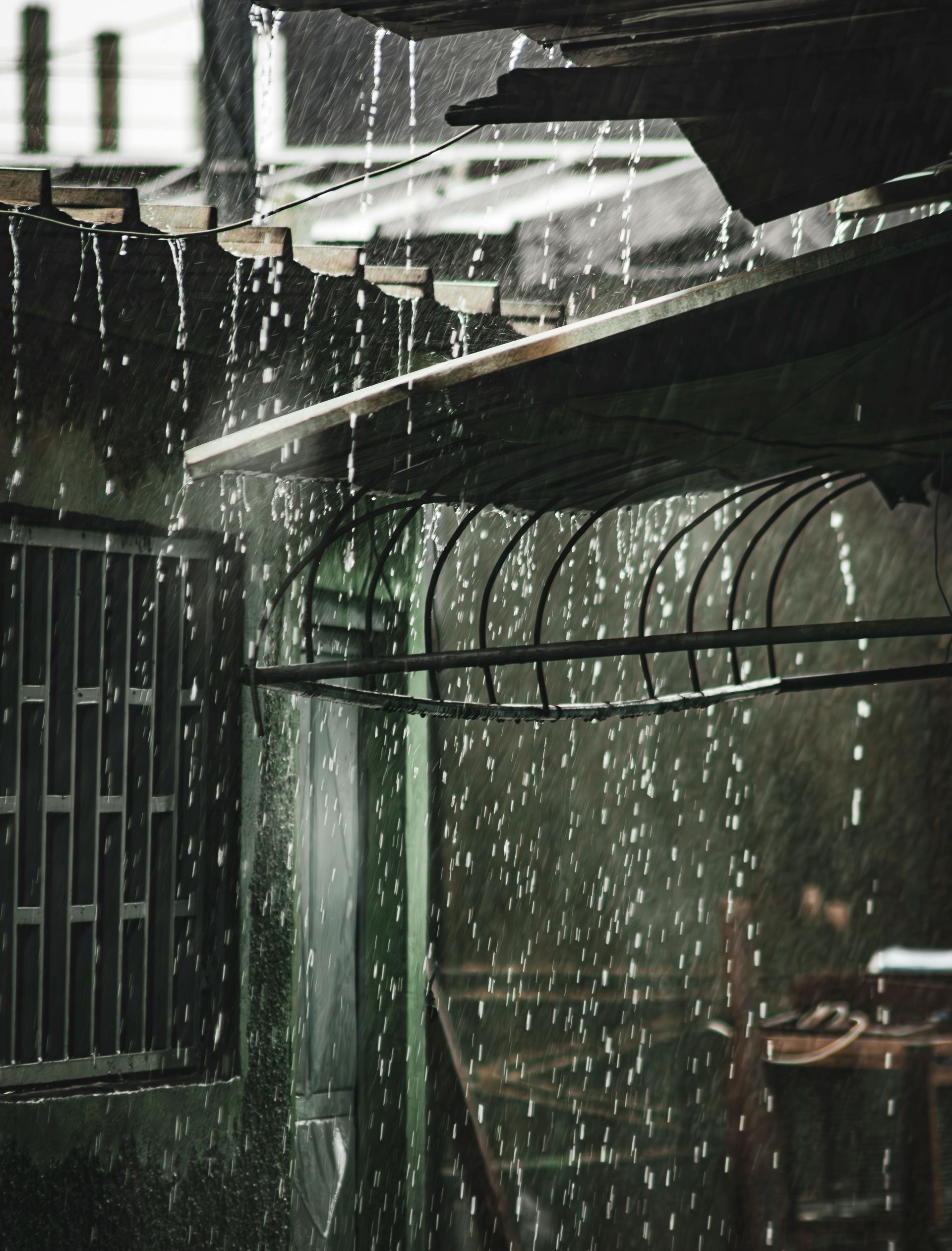 Heavy rain pours from the edge of a building roof, creating a blurred curtain of droplets over a dark, moody exterior.