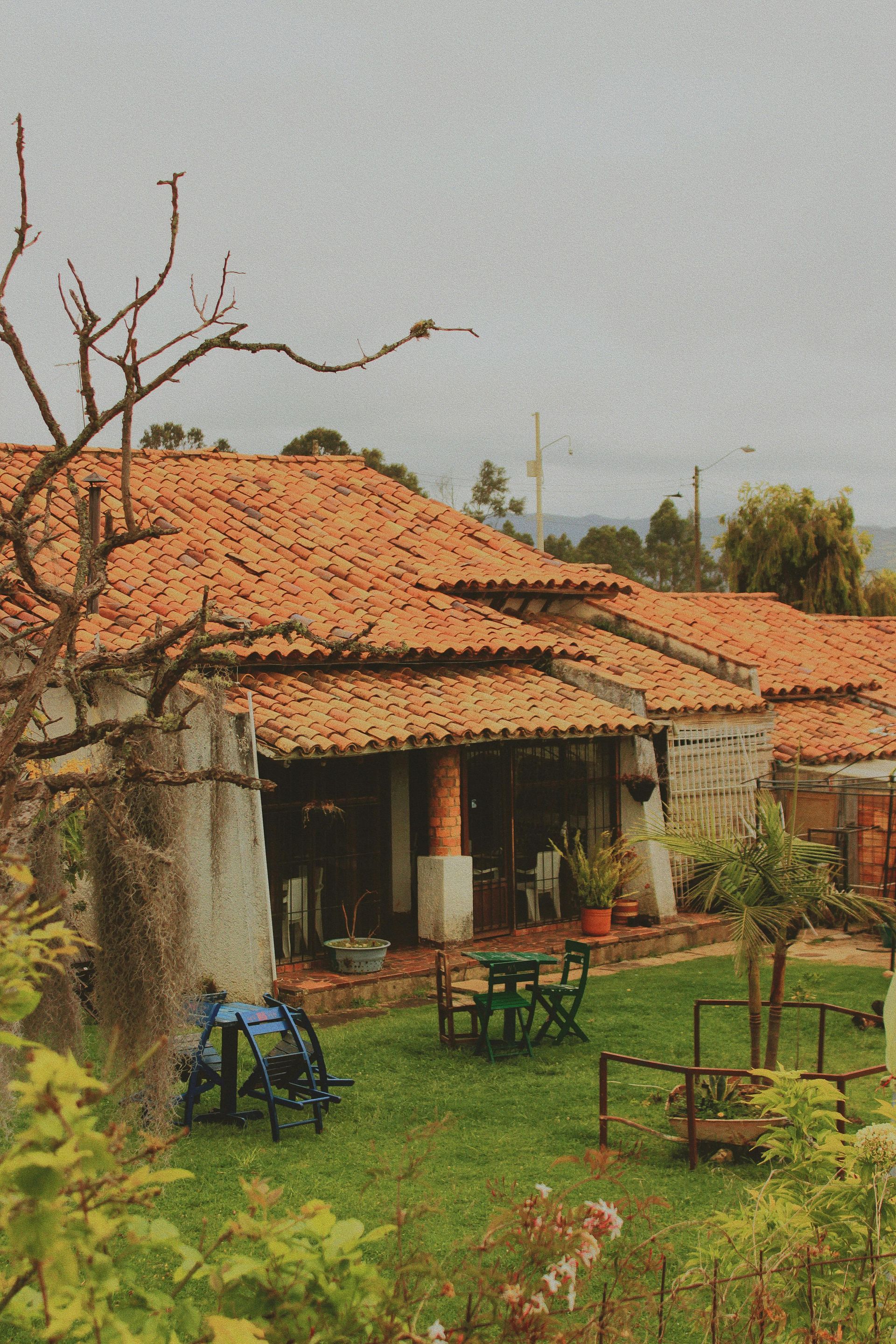 A cozy, rustic cottage with a terracotta tile roof, a lush green lawn, and patio furniture in a calm, outdoor setting.