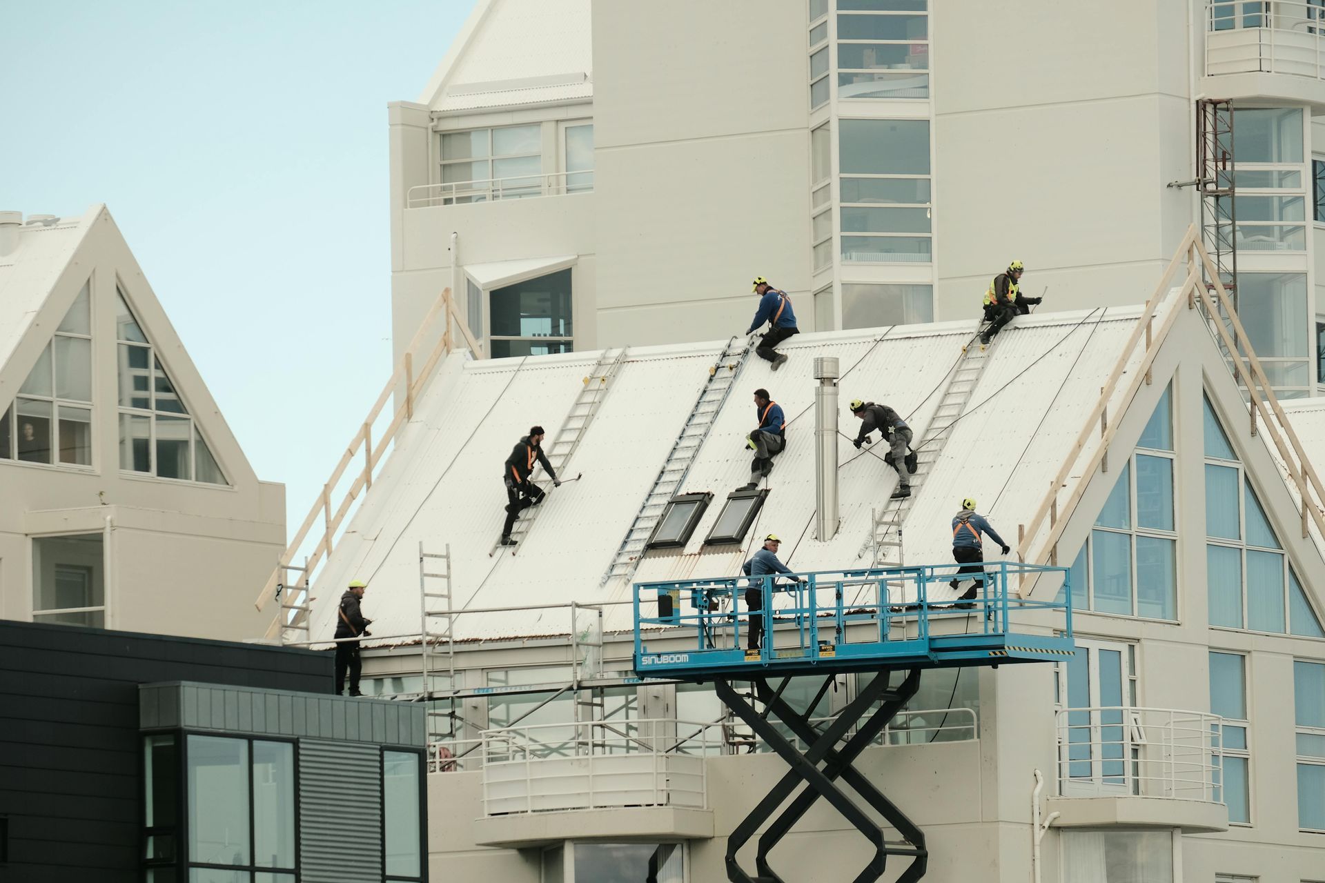 Workers on a sloping roof and a blue scissor lift, installing materials on a light-colored building.