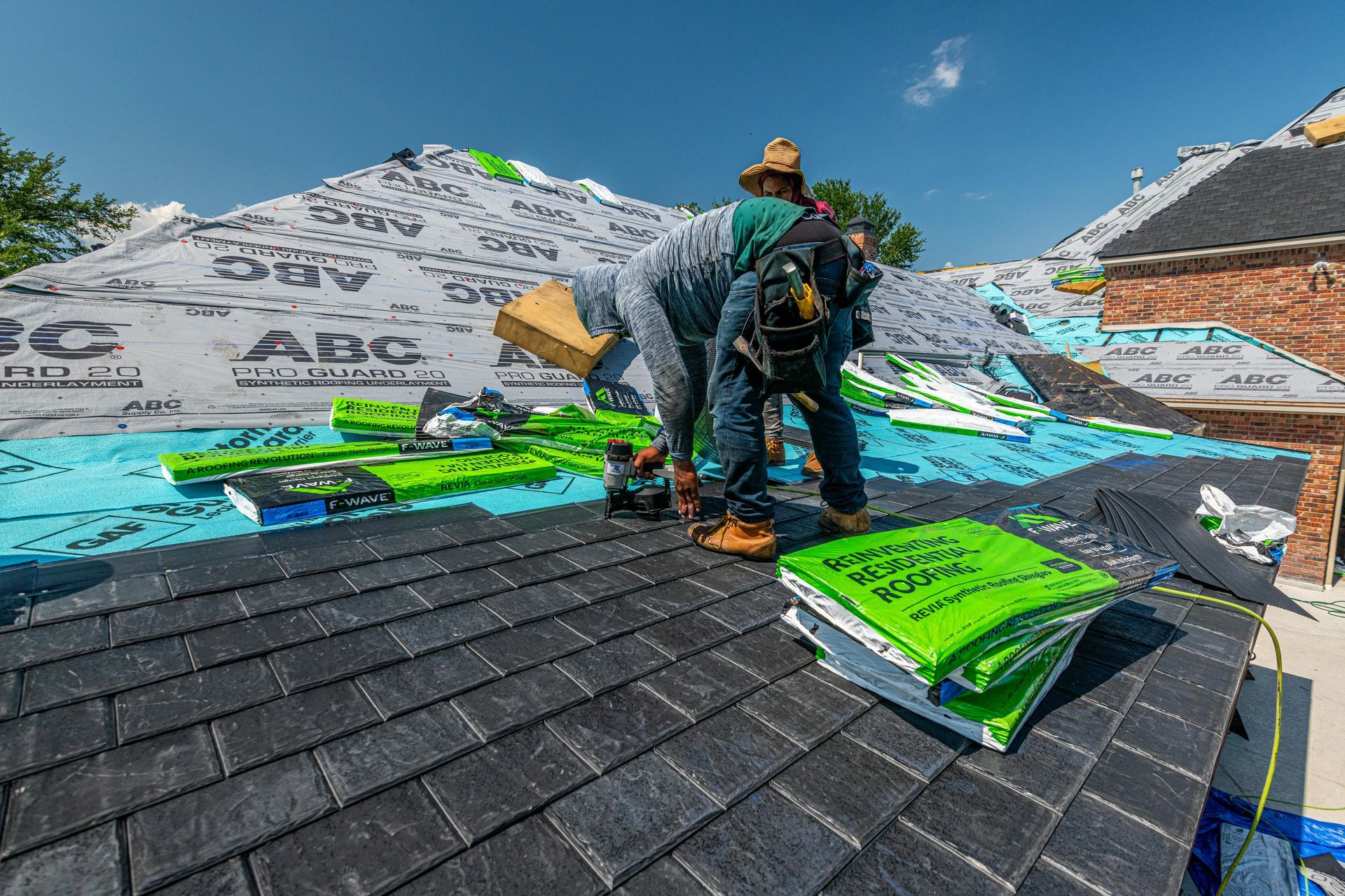 A roofer installs black shingles on a sloped roof with green building materials nearby under a clear blue sky.